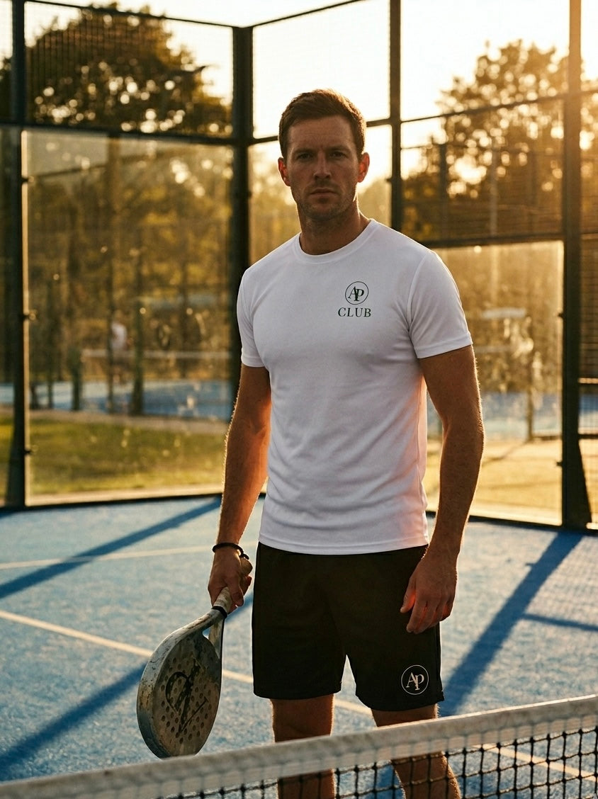 Man holding a padel racket on a padel court with trees in the background wearing a white AP Club Play Tee and black AP Club court shorts