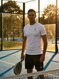 Man holding a padel racket on a padel court with trees in the background wearing a white AP Club Play Tee and black AP Club court shorts