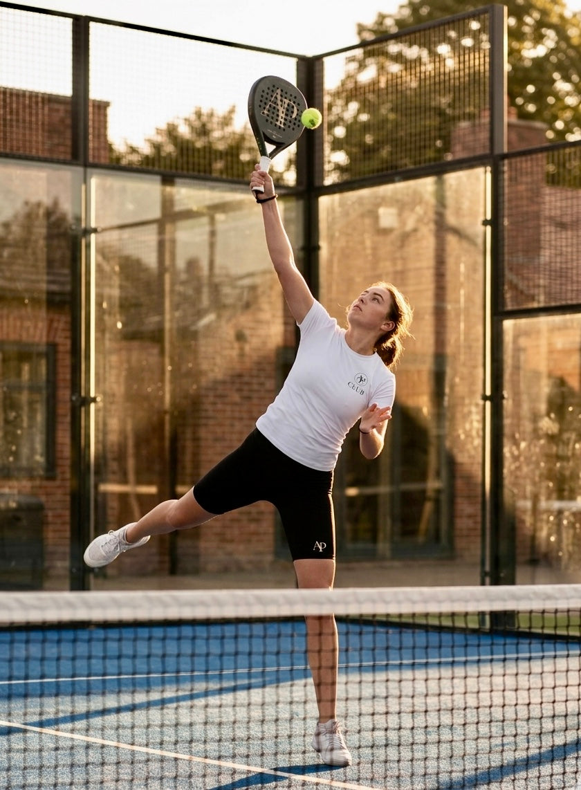 Woman playing padel tennis on a court with a glass wall background wearing a white AP Club Play Tee