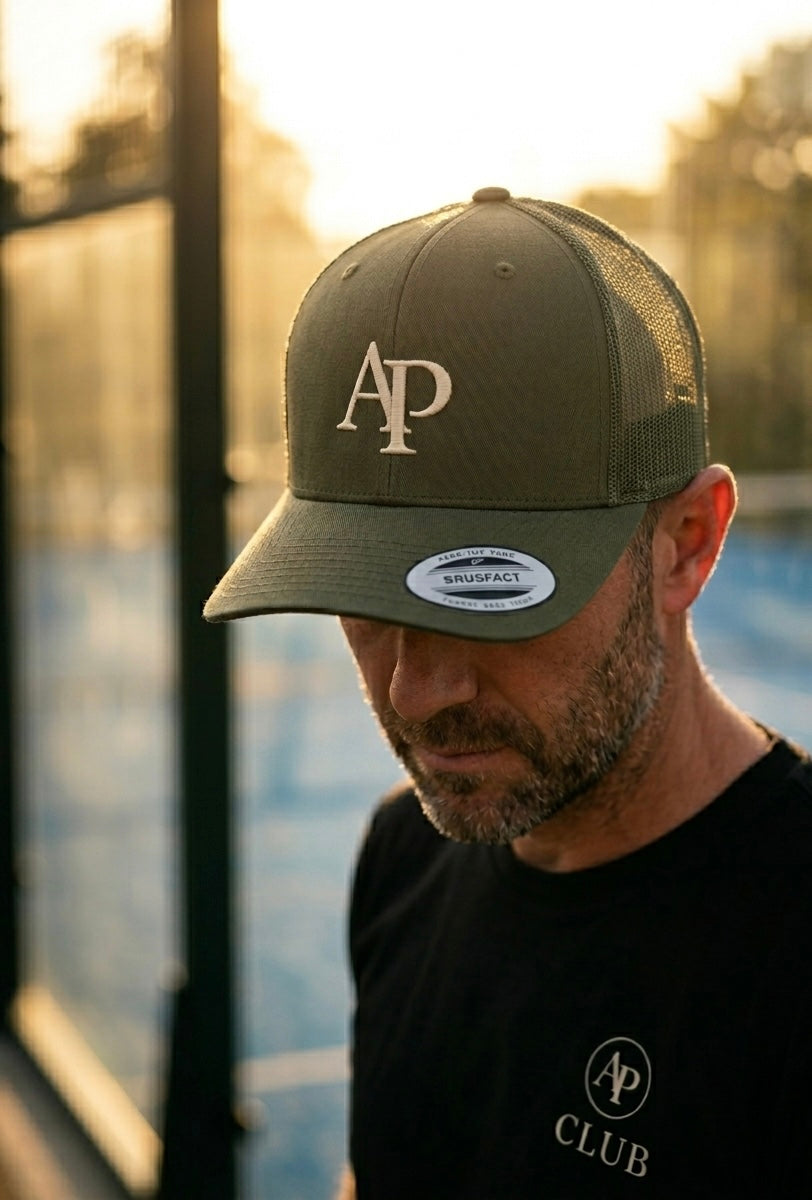 Man wearing a green AP Club cap with a logo, standing outdoors with a blurred background