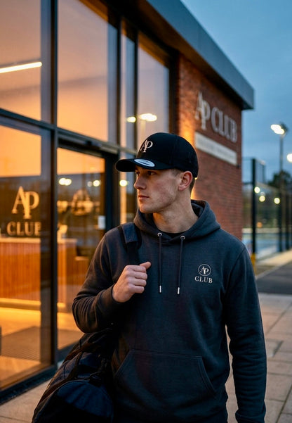 Man wearing a grey AP Club Vamos hoodie and AP Club cap with 'AP Club' branding in front of a padel court building.