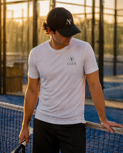 Man standing on a padel court holding a racket, wearing a white AP Club Play t-shirt and black AP Club cap with a logo.