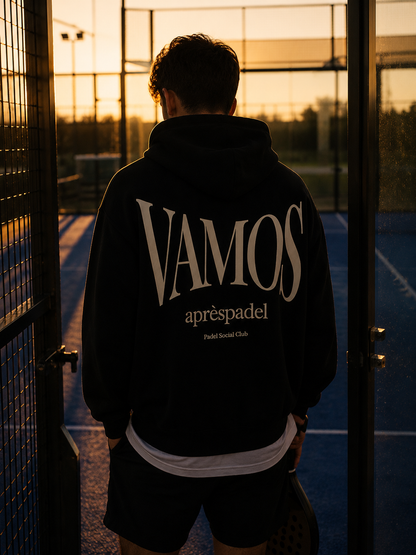 Person wearing a black AP Club VAMOS hoodie standing on a padel court at sunset.