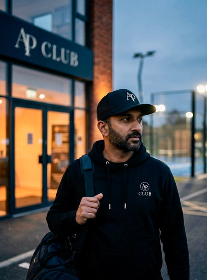Man wearing a black AP Club hoodie and AP Club cap with 'AP Club' branding, standing outside a padel court building.