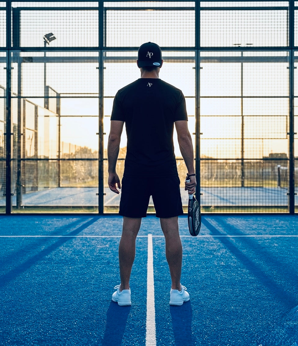 Person standing on a blue padel court holding a racket, wearing black AP Club play tee and Black AP Club cap.