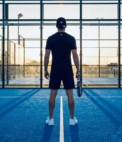 Person standing on a blue padel court holding a racket, wearing black AP Club play tee and Black AP Club cap.