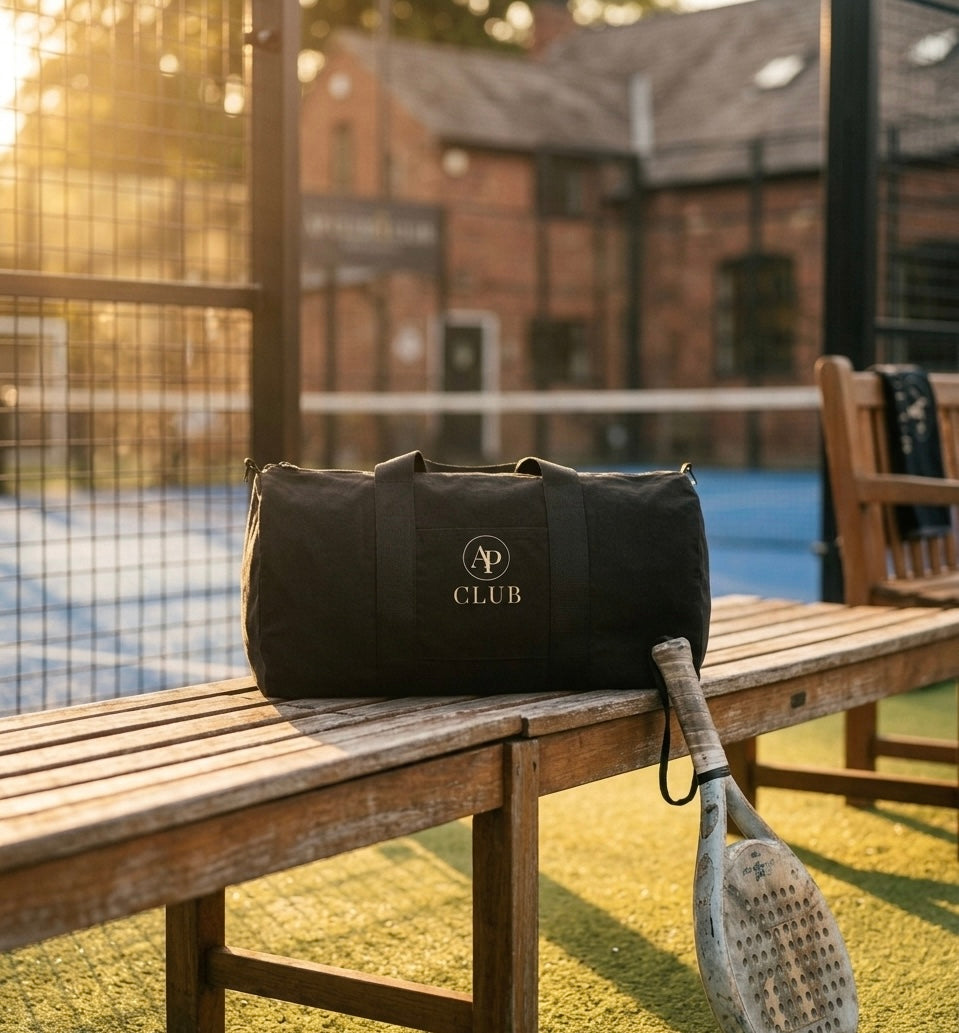 Black AP Club duffel bag with 'AP Club' logo on a wooden bench near a padel court.