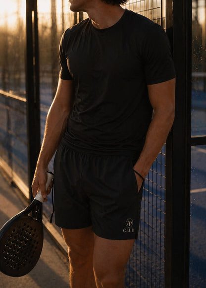 Man in black athletic AP Club Court shorts holding a paddle on a padel court at sunset.