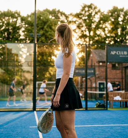 Woman holding a paddle on a padel court with other players in the background wearing a black AP club padel skort