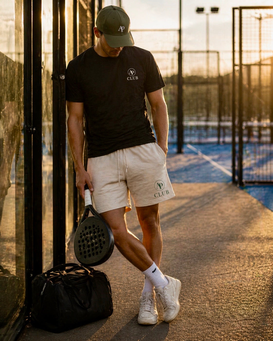 Man with a paddle racket and bag on a padel court at sunset wearing a black AP Club padel tshirt and AP Club club shorts AP Club cap