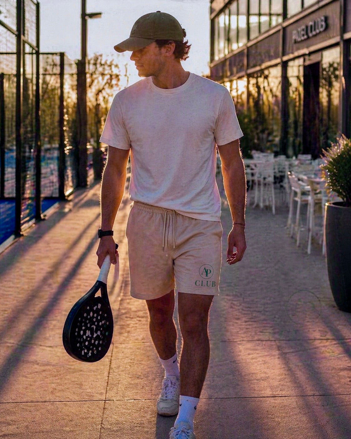 Man holding a paddle on a padel court with a building in the background wearing an AP Club cap