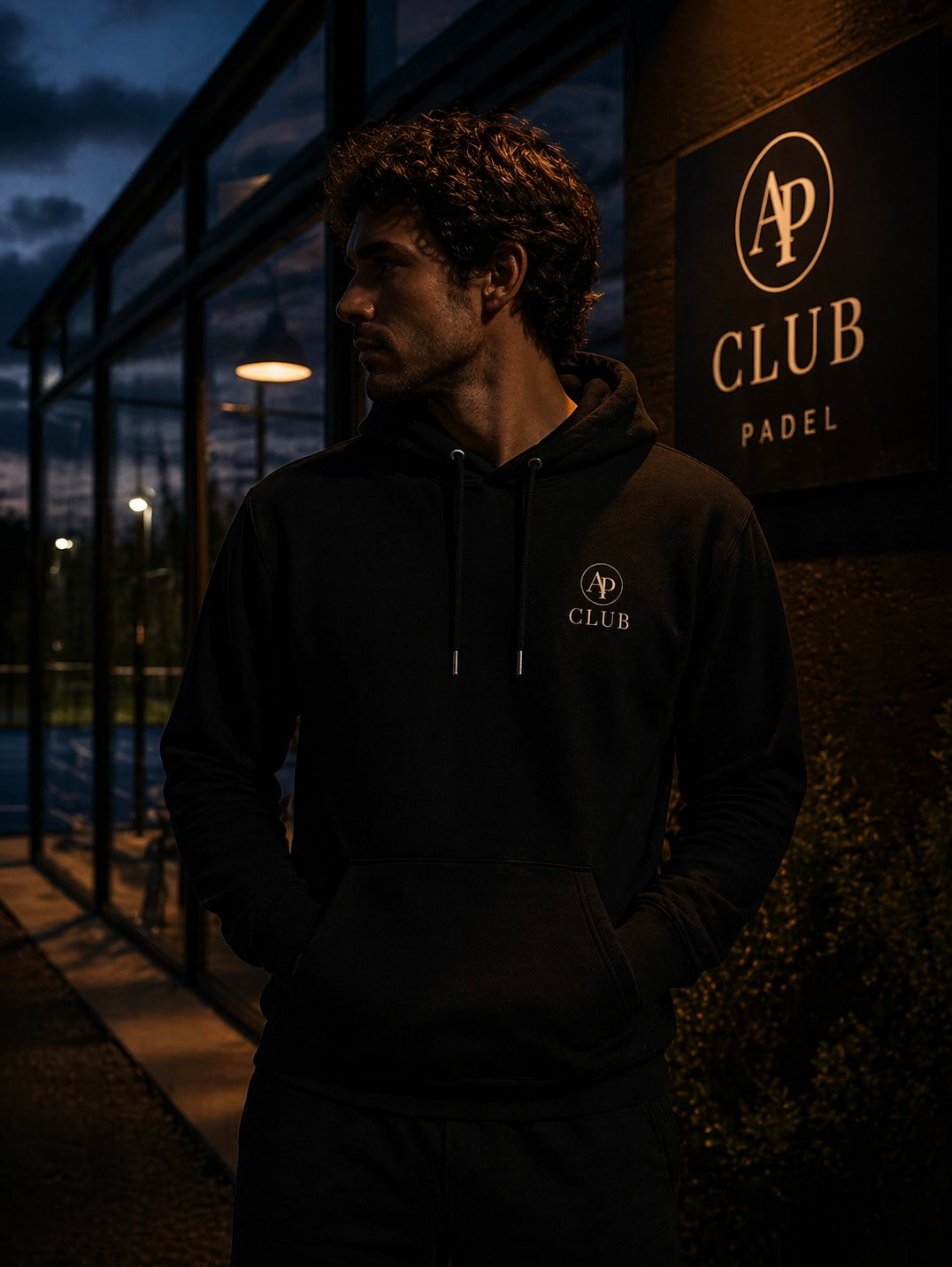 Person wearing a black AP Club Vamos hoodie with a logo standing in front of a building with 'AP Club Padel' sign.